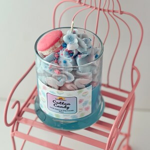 Puede incluir: Una vela de soja azul y rosa con aroma a algod&oacute;n de az&uacute;car. La vela est&aacute; en un tarro de vidrio con un macaron en la parte superior. El tarro est&aacute; sentado en una silla de metal rosa.