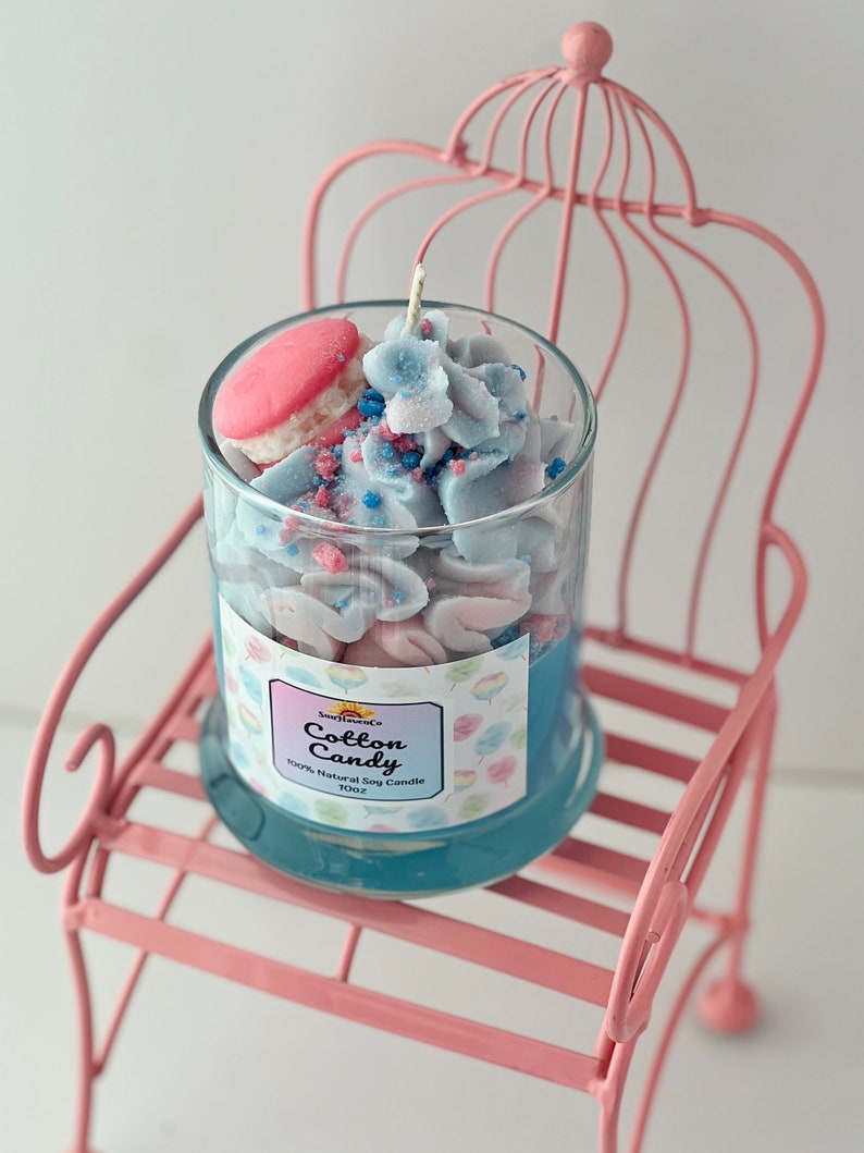 Puede incluir: Una vela de soja azul y rosa con aroma a algod&oacute;n de az&uacute;car. La vela est&aacute; en un tarro de vidrio con un macaron en la parte superior. El tarro est&aacute; sentado en una silla de metal rosa.