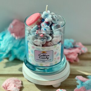 Puede incluir: Una vela de vidrio con aroma a algod&oacute;n de az&uacute;car azul y rosa. La vela est&aacute; cubierta con un macarr&oacute;n rosa, chispas azules y rosas y algod&oacute;n de az&uacute;car blanco. El frasco tiene una etiqueta que dice "SunHavenCo Cotton Candy 100% Natural Soy Candle 10oz".