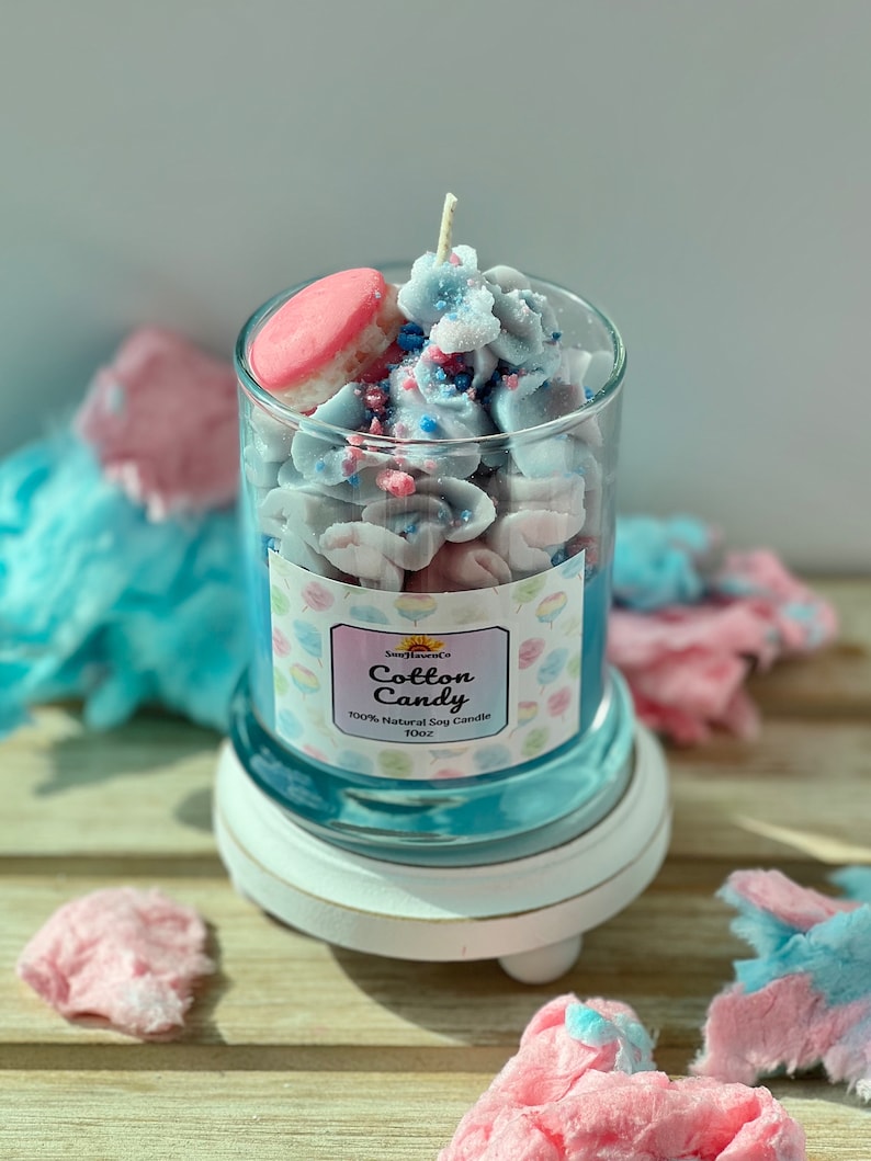 Puede incluir: Una vela de vidrio con aroma a algod&oacute;n de az&uacute;car azul y rosa. La vela est&aacute; cubierta con un macarr&oacute;n rosa, chispas azules y rosas y algod&oacute;n de az&uacute;car blanco. El frasco tiene una etiqueta que dice "SunHavenCo Cotton Candy 100% Natural Soy Candle 10oz".