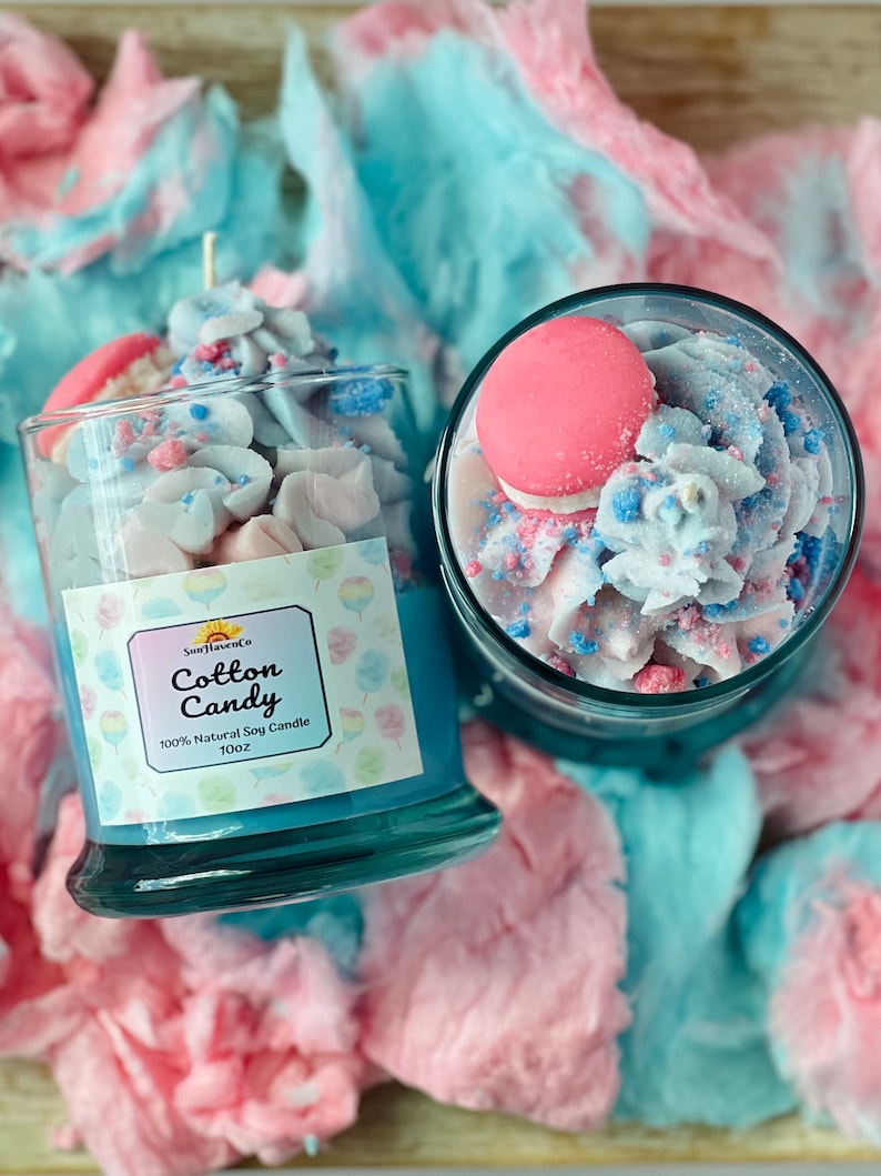 Puede incluir: Dos frascos de vidrio llenos de una vela de soja con aroma a algod&oacute;n de az&uacute;car azul y rosa. Los frascos est&aacute;n rodeados de algod&oacute;n de az&uacute;car rosa y azul. La etiqueta del frasco dice "Cotton Candy 100% Natural Soy Candle 10oz"