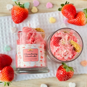 May include: A pink strawberry shortcake scented candle in a glass jar with a white background. The candle is decorated with a pink and white frosting design and a small strawberry on top. The jar has a label that says "Strawberry Shortcake" and "100% Natural Soy Candle 10oz".