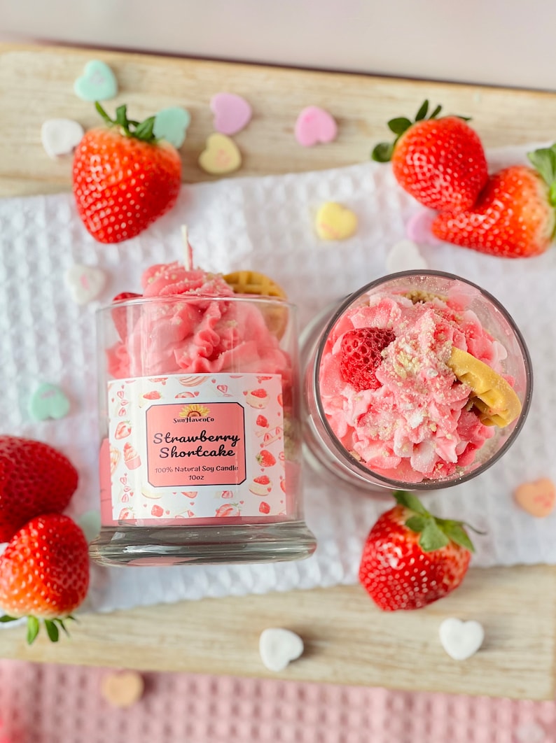 May include: A pink strawberry shortcake scented candle in a glass jar with a white background. The candle is decorated with a pink and white frosting design and a small strawberry on top. The jar has a label that says "Strawberry Shortcake" and "100% Natural Soy Candle 10oz".