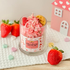 May include: A pink and white strawberry shortcake scented soy candle in a glass jar with a waffle and strawberry on top. The candle is sitting on a white crocheted surface with pink and white heart-shaped candies.