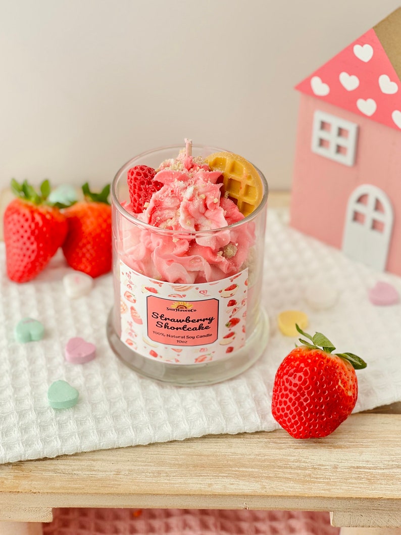 May include: A pink and white strawberry shortcake scented soy candle in a glass jar with a waffle and strawberry on top. The candle is sitting on a white crocheted surface with pink and white heart-shaped candies.