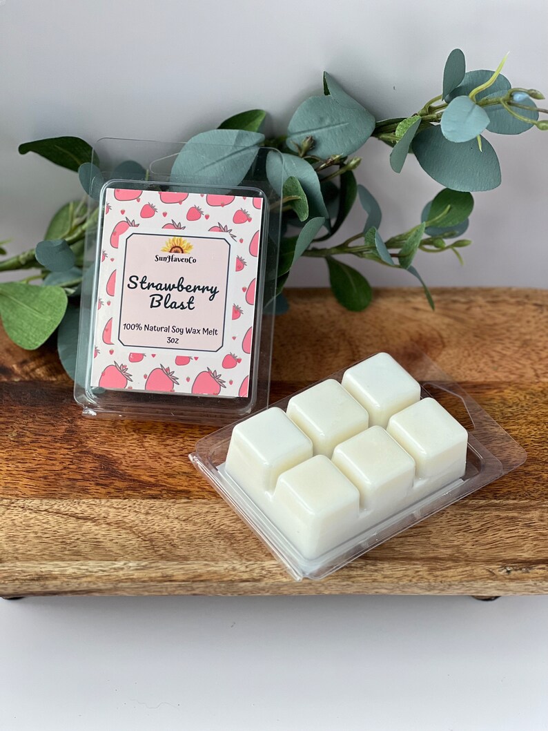 May include: Six white wax melts in a clear plastic tray. The tray is on a wooden surface. A smaller clear plastic tray with a label that reads "Strawberry Blast" and "SunHavenCo" is behind the wax melts. The label has a pink and white background with a strawberry pattern.