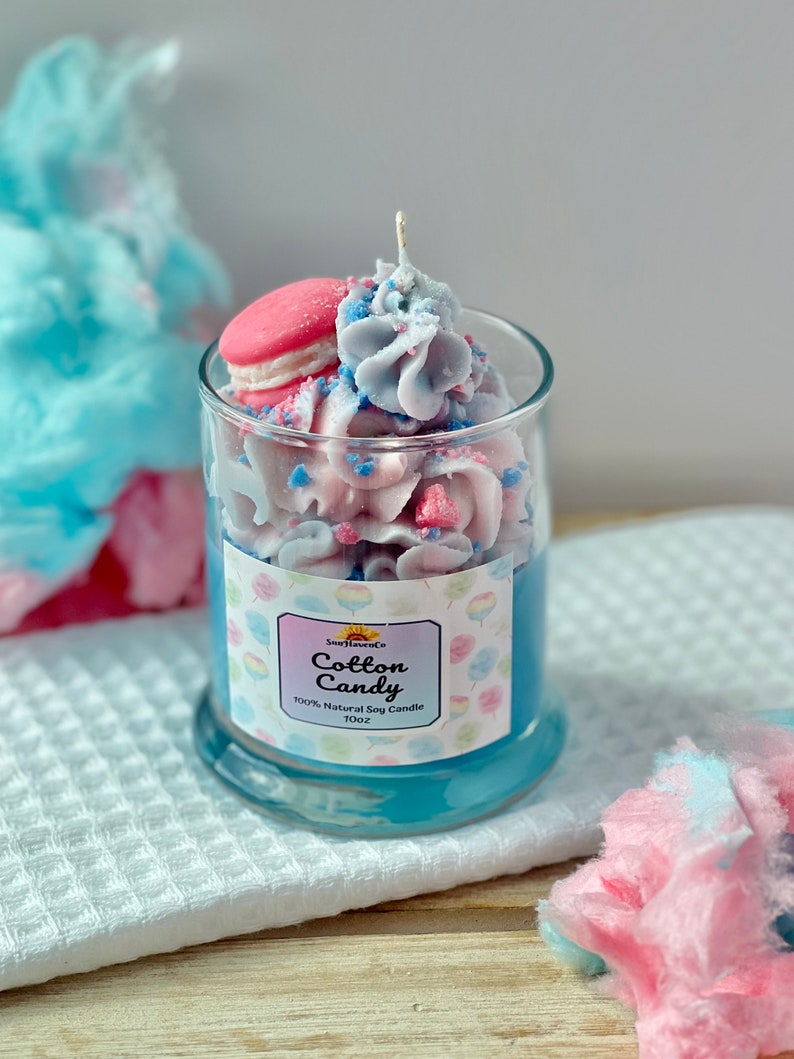 Puede incluir: Una vela de vidrio azul con aroma a algod&oacute;n de az&uacute;car. La vela est&aacute; cubierta con un macarr&oacute;n rosa, glaseado azul y rosa, y chispas azules y rosas. El frasco tiene una etiqueta que dice "SunhavenCo Cotton Candy 100% Natural Soy Candle 10oz".