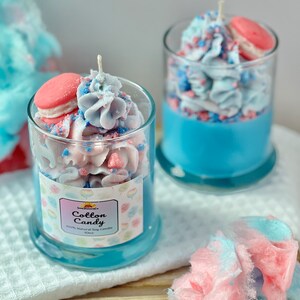 Puede incluir: Dos frascos de vidrio llenos de velas arom&aacute;ticas de algod&oacute;n de az&uacute;car azul y rosa. Las velas est&aacute;n coronadas con un macarr&oacute;n rosa y glaseado blanco. Los frascos tienen una etiqueta que dice "Cotton Candy" y "100% Natural Soy Candle 10oz".