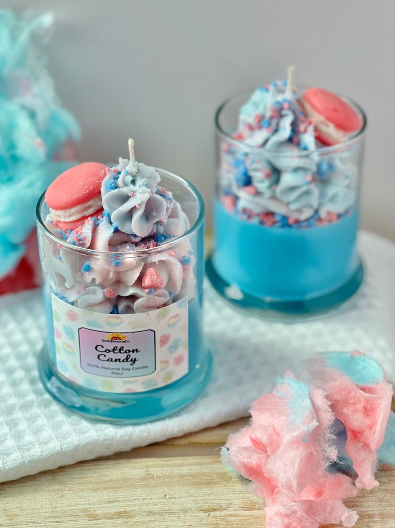 Puede incluir: Dos frascos de vidrio llenos de velas arom&aacute;ticas de algod&oacute;n de az&uacute;car azul y rosa. Las velas est&aacute;n coronadas con un macarr&oacute;n rosa y glaseado blanco. Los frascos tienen una etiqueta que dice "Cotton Candy" y "100% Natural Soy Candle 10oz".