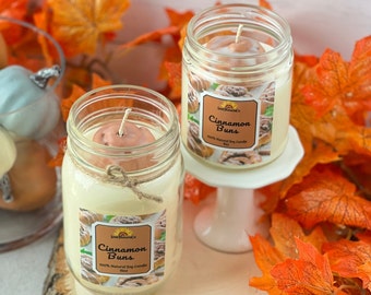 May include: Two glass jars with lids containing soy candles. The candles are labeled "Cinnamon Buns" and are 100% natural soy candles. The jars are sitting on a wooden surface with fall leaves around them.