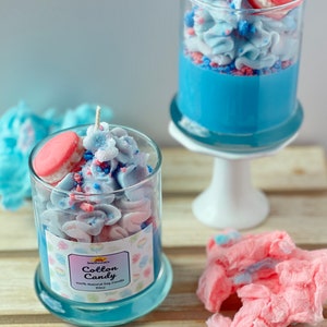 Puede incluir: Dos tarros de vidrio llenos de velas arom&aacute;ticas de algod&oacute;n de az&uacute;car azul y rosa. Las velas est&aacute;n coronadas con un macarr&oacute;n rosa y espolvoreadas con chispas azules y rosas. Los tarros est&aacute;n sobre una superficie de madera. El texto de la etiqueta dice "Cotton Candy 100% Natural Soy Candle 10oz".