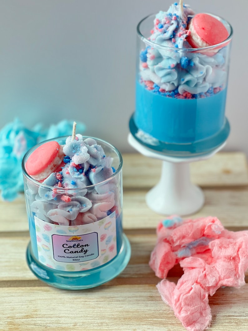 Puede incluir: Dos tarros de vidrio llenos de velas arom&aacute;ticas de algod&oacute;n de az&uacute;car azul y rosa. Las velas est&aacute;n coronadas con un macarr&oacute;n rosa y espolvoreadas con chispas azules y rosas. Los tarros est&aacute;n sobre una superficie de madera. El texto de la etiqueta dice "Cotton Candy 100% Natural Soy Candle 10oz".