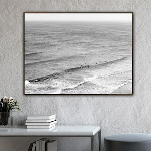 以下が含まれることがあります： サーファーがいる海の波の白黒プリント。アートワークは、本と花瓶のある白いテーブルの上にあります。灰色のオットマンが近くにあります。部屋にはレンガの壁と大きな窓があります。