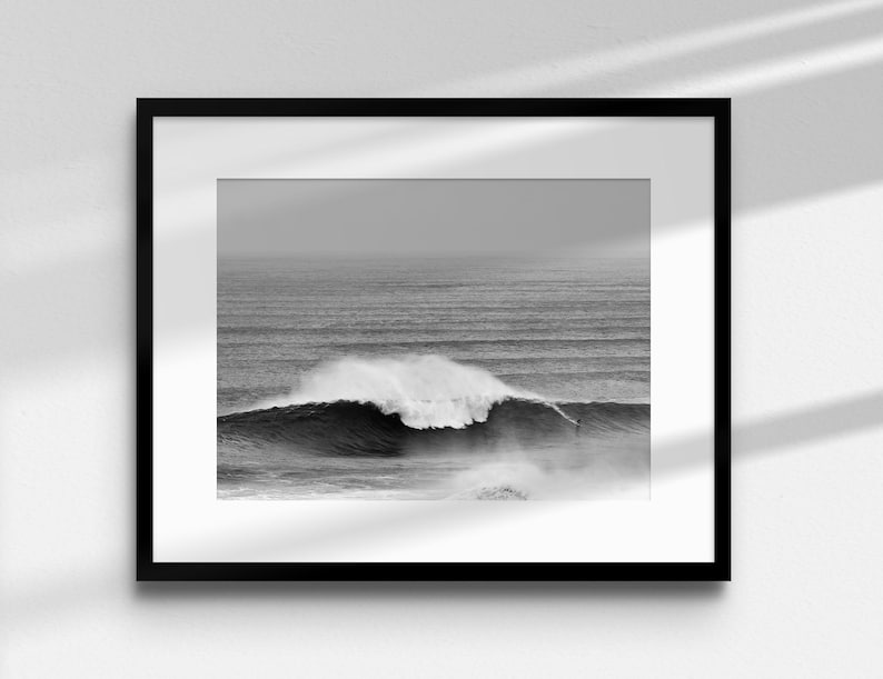 Monochrome ocean wall art featuring a big wave surfer in Nazaré. The image is framed with a white mat and displayed in a neutral-toned room. Perfect for lovers of surfing, minimalism, and Atlantic landscapes. High-quality digital file for large format printing.