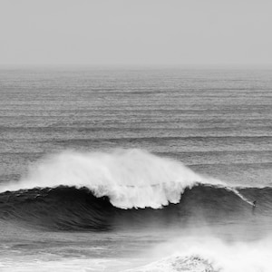 Digital black and white surf print of a giant wave in Portugal with a small surfer riding its face. Captured in Nazaré. High-resolution photo for framing, coastal wall decor, or modern gallery walls. Ideal for lovers of scale, movement, and the ocean.