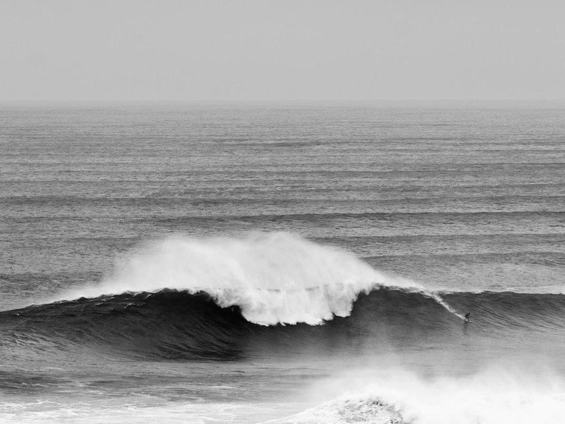 Digital black and white surf print of a giant wave in Portugal with a small surfer riding its face. Captured in Nazaré. High-resolution photo for framing, coastal wall decor, or modern gallery walls. Ideal for lovers of scale, movement, and the ocean.