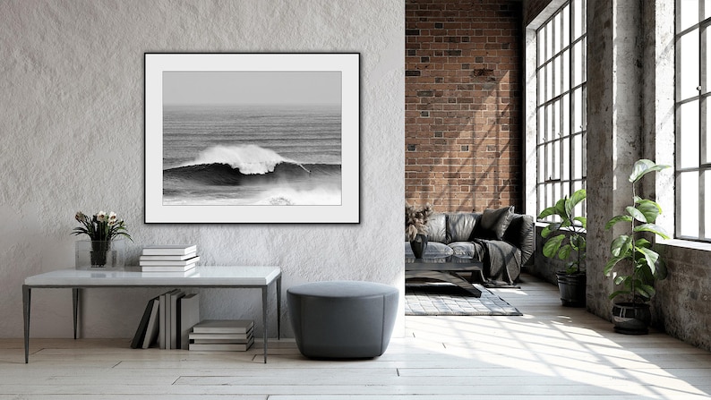 Striking black and white print of a big wave surfer in Nazaré, Portugal. Hung in an industrial loft living room with raw concrete and brick walls. A dramatic, inspiring ocean photo — ideal for modern masculine or creative interiors.
