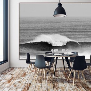 Moody black and white surfing print displayed in a minimalist dining room. The photograph shows a big wave in Nazaré with a surfer dwarfed by the scale. Coastal digital download art for serene, textured interiors with a bold twist.