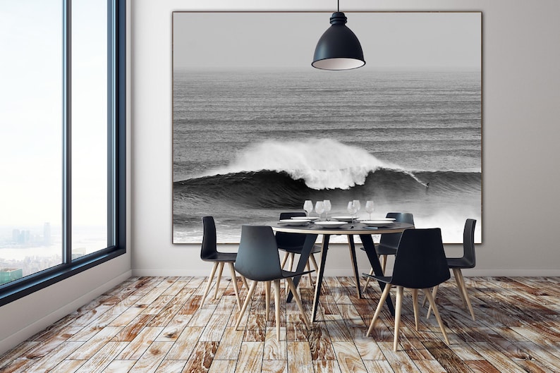 Moody black and white surfing print displayed in a minimalist dining room. The photograph shows a big wave in Nazaré with a surfer dwarfed by the scale. Coastal digital download art for serene, textured interiors with a bold twist.
