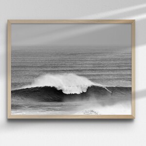 Black and white surf photography print showing a surfer on a giant wave in Nazaré, Portugal. Framed in minimalist black wood and styled in a modern living space. Powerful ocean scene for coastal, Japandi, or loft interiors. High-resolution digital download artwork.