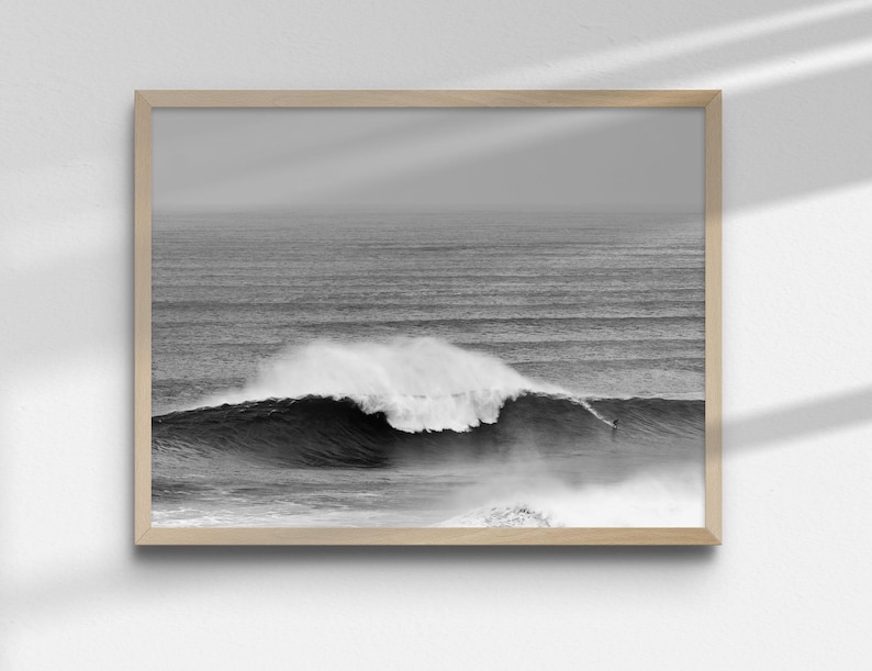 Black and white surf photography print showing a surfer on a giant wave in Nazaré, Portugal. Framed in minimalist black wood and styled in a modern living space. Powerful ocean scene for coastal, Japandi, or loft interiors. High-resolution digital download artwork.