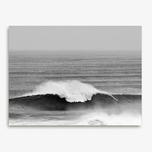 Digital black and white surf print of a giant wave in Portugal with a small surfer riding its face. Captured in Nazaré. High-resolution photo for framing, coastal wall decor, or modern gallery walls. Ideal for lovers of scale, movement, and the ocean.