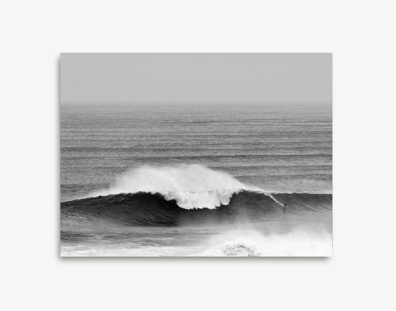 Digital black and white surf print of a giant wave in Portugal with a small surfer riding its face. Captured in Nazaré. High-resolution photo for framing, coastal wall decor, or modern gallery walls. Ideal for lovers of scale, movement, and the ocean.