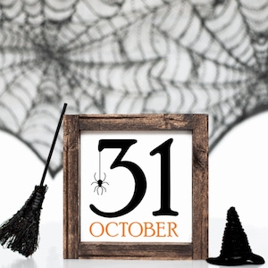May include: A Halloween-themed wooden sign with the number 31 and the word "October" in orange. A small black spider hangs from the number 31. A black witch's hat and broom are also present, set against a backdrop of a spider web.