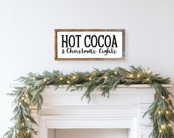 May include: A wooden sign with the text "HOT COCOA & Christmas lights" in black lettering on a white background. The sign is hanging above a white fireplace mantel decorated with a green garland with white lights.