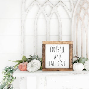 May include: A wooden framed sign with the text "FOOTBALL AND FALL Y'ALL" sits on a white shelf, decorated with pumpkins and greenery. The sign is surrounded by white and orange pumpkins, and green foliage.
