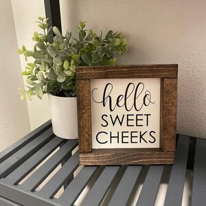 Peut inclure: Une petite pancarte en bois avec un fond blanc et des lettres noires qui disent "hello sweet cheeks". La pancarte est pos&eacute;e sur une &eacute;tag&egrave;re &agrave; lattes grises.