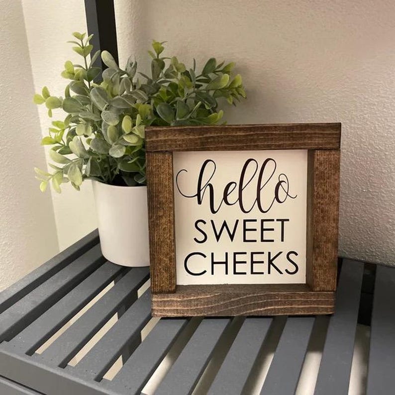 Peut inclure: Une petite pancarte en bois avec un fond blanc et des lettres noires qui disent "hello sweet cheeks". La pancarte est pos&eacute;e sur une &eacute;tag&egrave;re &agrave; lattes grises.
