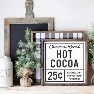 May include: A white sign with black text that reads "Christmas Blend Hot Cocoa 25&cent; Marshmallows Whipped Cream Peppermint". The sign is framed in wood and is sitting on a shelf with a white ceramic coffee canister and two small artificial pine trees.