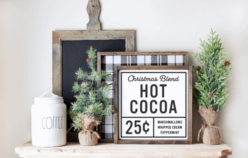 May include: A white sign with black text that reads "Christmas Blend Hot Cocoa 25&cent; Marshmallows Whipped Cream Peppermint". The sign is framed in wood and is sitting on a shelf with a white ceramic coffee canister and two small artificial pine trees.