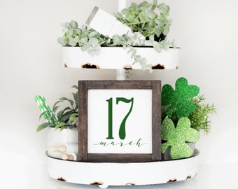 May include: A white tiered tray with green plants and a wooden sign that says "17 march". The sign has a green number 17 and the word "march" is written in cursive.