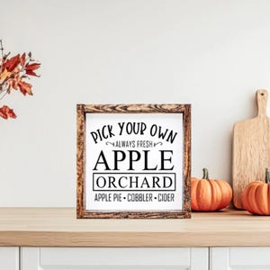 May include: A wooden framed sign with the text "Pick Your Own Apple Orchard" and the words "Apple Pie, Cobbler, Cider". The sign is on a wooden surface with pumpkins, a cutting board, and a vase of fall foliage.