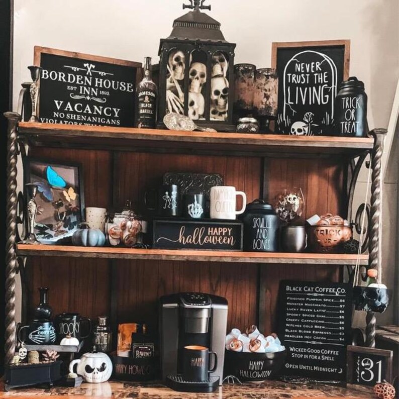 May include: A Halloween display on a wooden shelf, featuring skulls, mugs, and signs with phrases like "Happy Halloween" and "Trick or Treat." There's also a coffee machine. The colour scheme is mainly black, white, and brown, creating a spooky atmosphere.