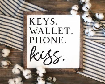 May include: White wooden sign with brown wood frame. The sign says "KEYS. WALLET. PHONE. kiss."