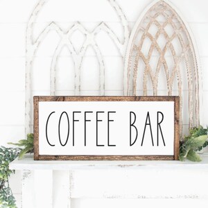 May include: A rectangular wooden sign with the words "COFFEE BAR" in black lettering on a white background. The sign is framed in a dark brown wood and is displayed on a white surface with decorative greenery.