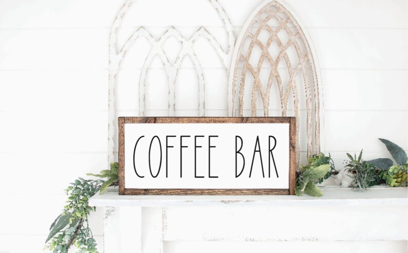 May include: A rectangular wooden sign with the words "COFFEE BAR" in black lettering on a white background. The sign is framed in a dark brown wood and is displayed on a white surface with decorative greenery.