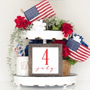 May include: A tiered tray decorated for the Fourth of July with red, white and blue American flags, red flowers, and a sign that says "4 July".