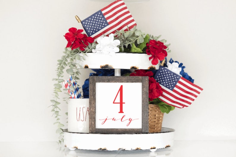 May include: A tiered tray decorated for the Fourth of July with red, white and blue American flags, red flowers, and a sign that says "4 July".