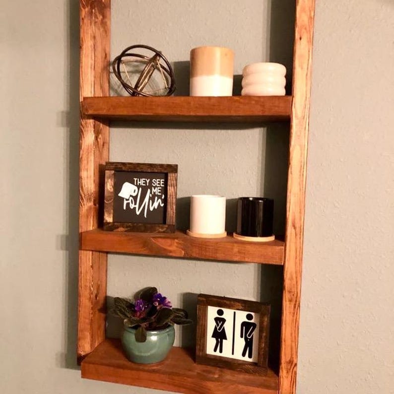 May include: A wooden ladder shelf with three shelves. The top shelf has a metal orb, a white and brown planter, and a white planter with a textured design. The middle shelf has a black framed sign that says "They see me rollin'" and two small planters, one white and one black. The bottom shelf has a small green planter with purple flowers and a black framed sign with a male and female restroom symbol.