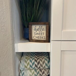 Peut inclure: Une petite pancarte en bois avec le texte "hello sweet cheeks" en lettres noires sur fond blanc. La pancarte est pos&eacute;e sur une &eacute;tag&egrave;re blanche avec une plante verte dans un pot bleu derri&egrave;re elle.