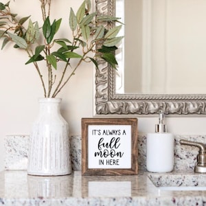 May include: A wooden sign with the text "It's always a full moon in here" in black lettering. The sign is sitting on a white countertop with a white vase and a white soap dispenser.