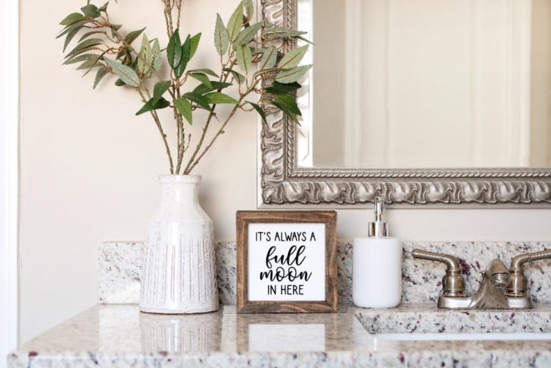 May include: A wooden sign with the text "It's always a full moon in here" in black lettering. The sign is sitting on a white countertop with a white vase and a white soap dispenser.