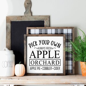 May include: A rustic shelf displays a white jar labeled "COFFEE", a small pumpkin, and a framed sign that reads "PICK YOUR OWN APPLE ORCHARD". A fern in an orange pot and a chalkboard complete the fall-themed decor.