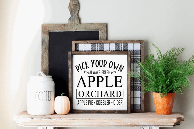 May include: A rustic shelf displays a white jar labeled "COFFEE", a small pumpkin, and a framed sign that reads "PICK YOUR OWN APPLE ORCHARD". A fern in an orange pot and a chalkboard complete the fall-themed decor.