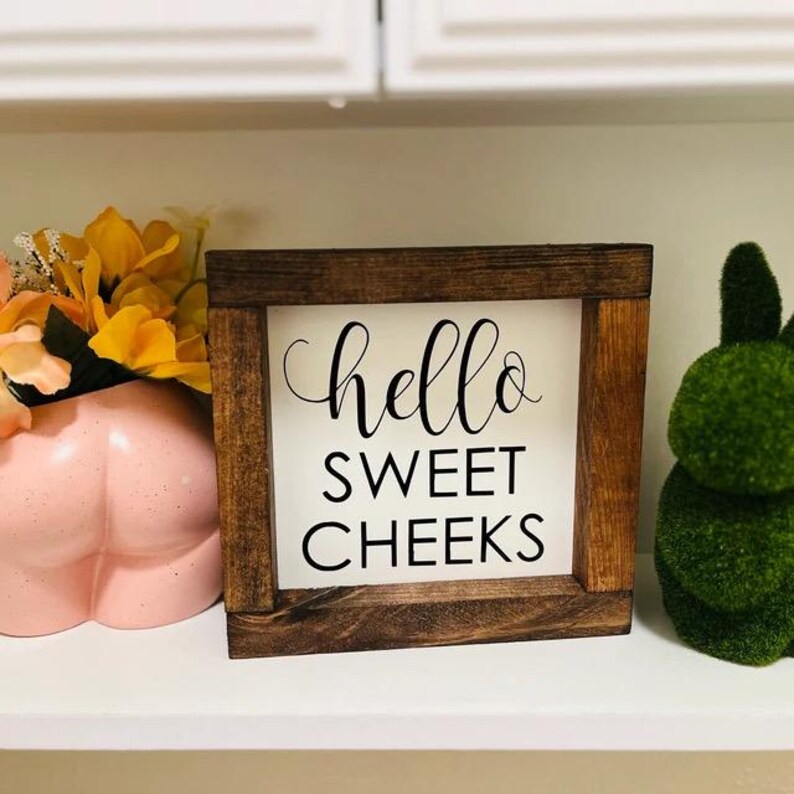 Peut inclure: Une pancarte en bois avec le texte "hello sweet cheeks" en lettres noires sur fond blanc. La pancarte est pos&eacute;e sur une &eacute;tag&egrave;re avec une figurine en c&eacute;ramique rose et une figurine de lapin en mousse verte.