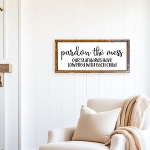 May include: A wooden sign with the text "pardon the mess our standards have lowered with each child" in black lettering.